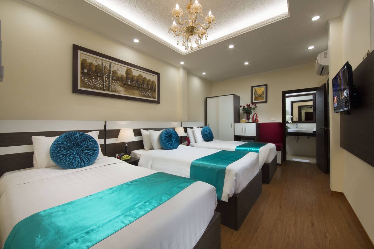 A hotel room with three beds, a chandelier, a painting, a mirror, and a bathroom.