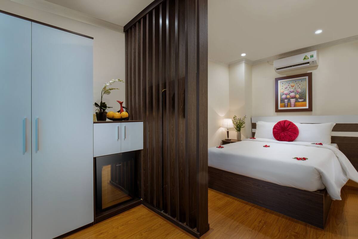 A modern hotel room with a wooden wardrobe, a bed with white bedding and a red pillow, and a painting on the wall.