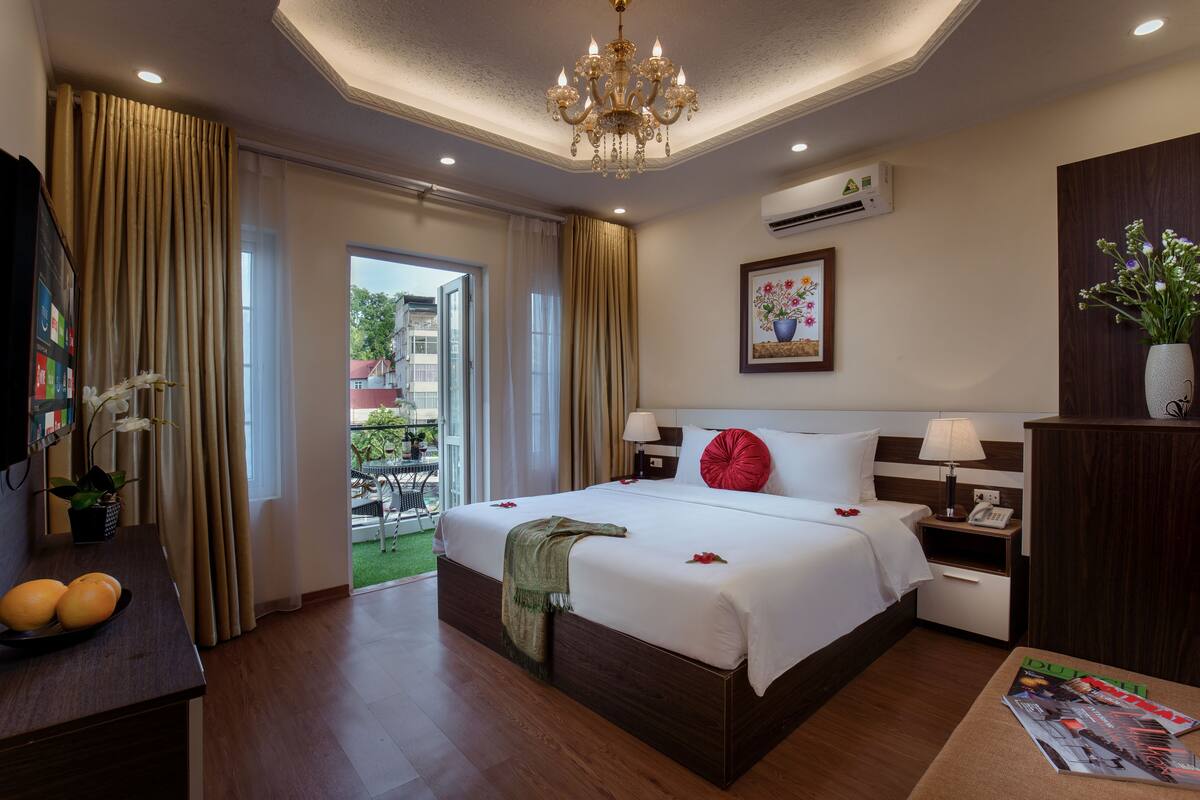 A hotel room with a large bed, a desk with a computer, a chair, a window with curtains, and a chandelier.