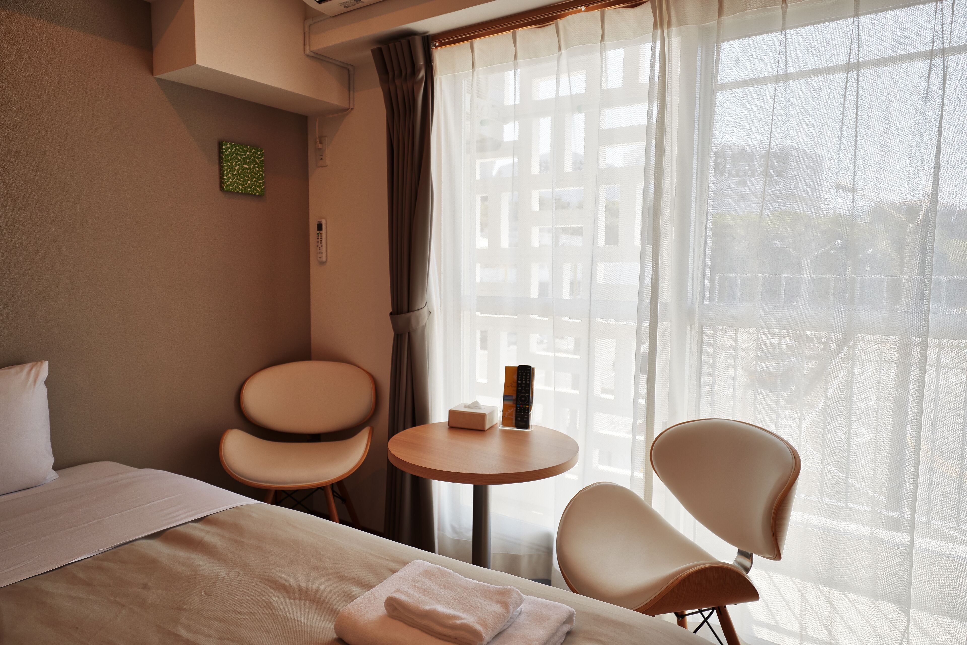 Habitación de hotel con cama, dos sillas, una mesita redonda y una ventana con cortinas traslúcidas.