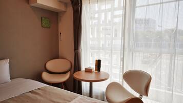 Habitación de hotel con cama, dos sillas, una mesita redonda y una ventana con cortinas traslúcidas.