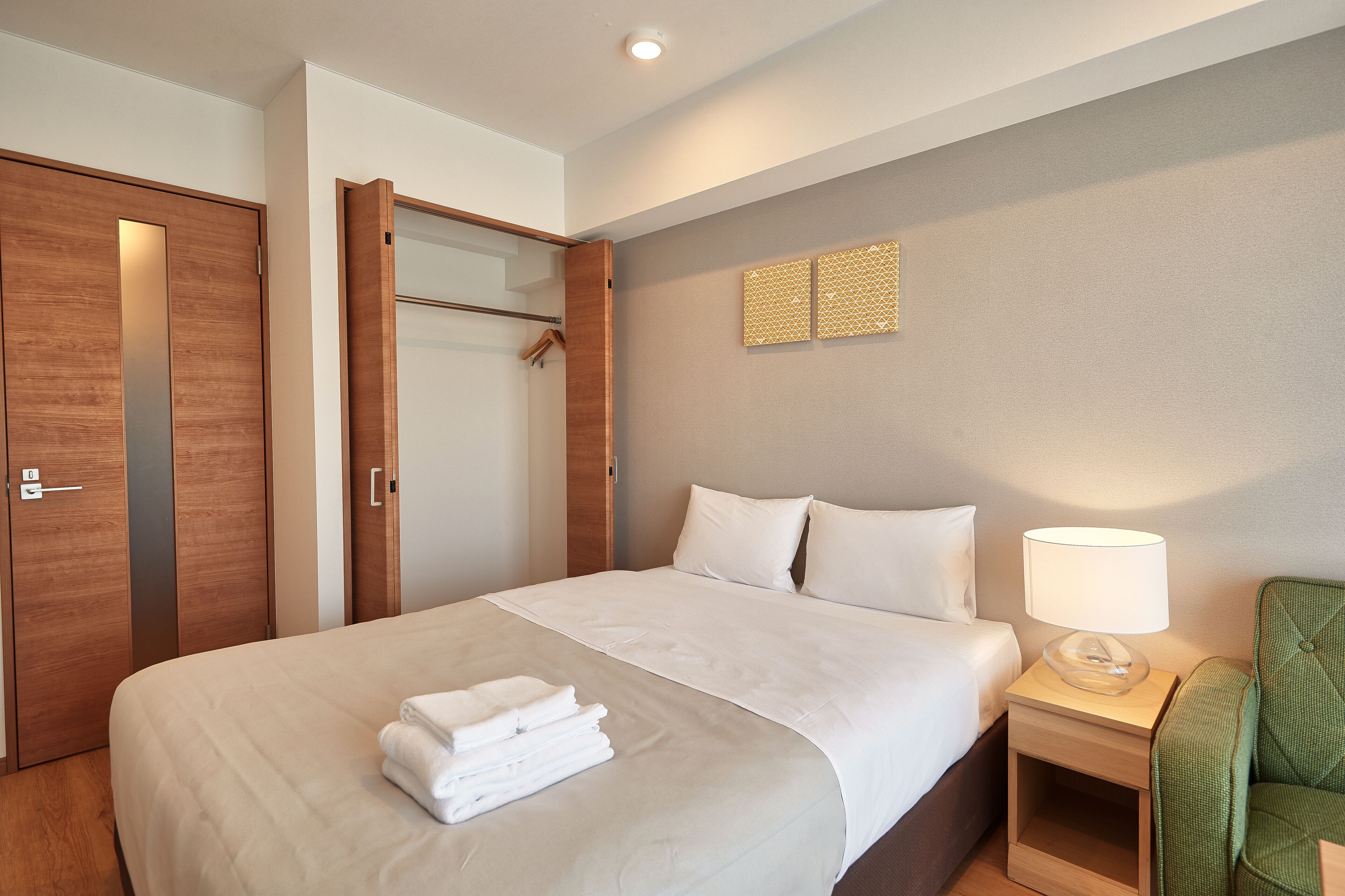 Una camera d'albergo con un letto, un comodino con una lampada, una sedia verde e un armadio.