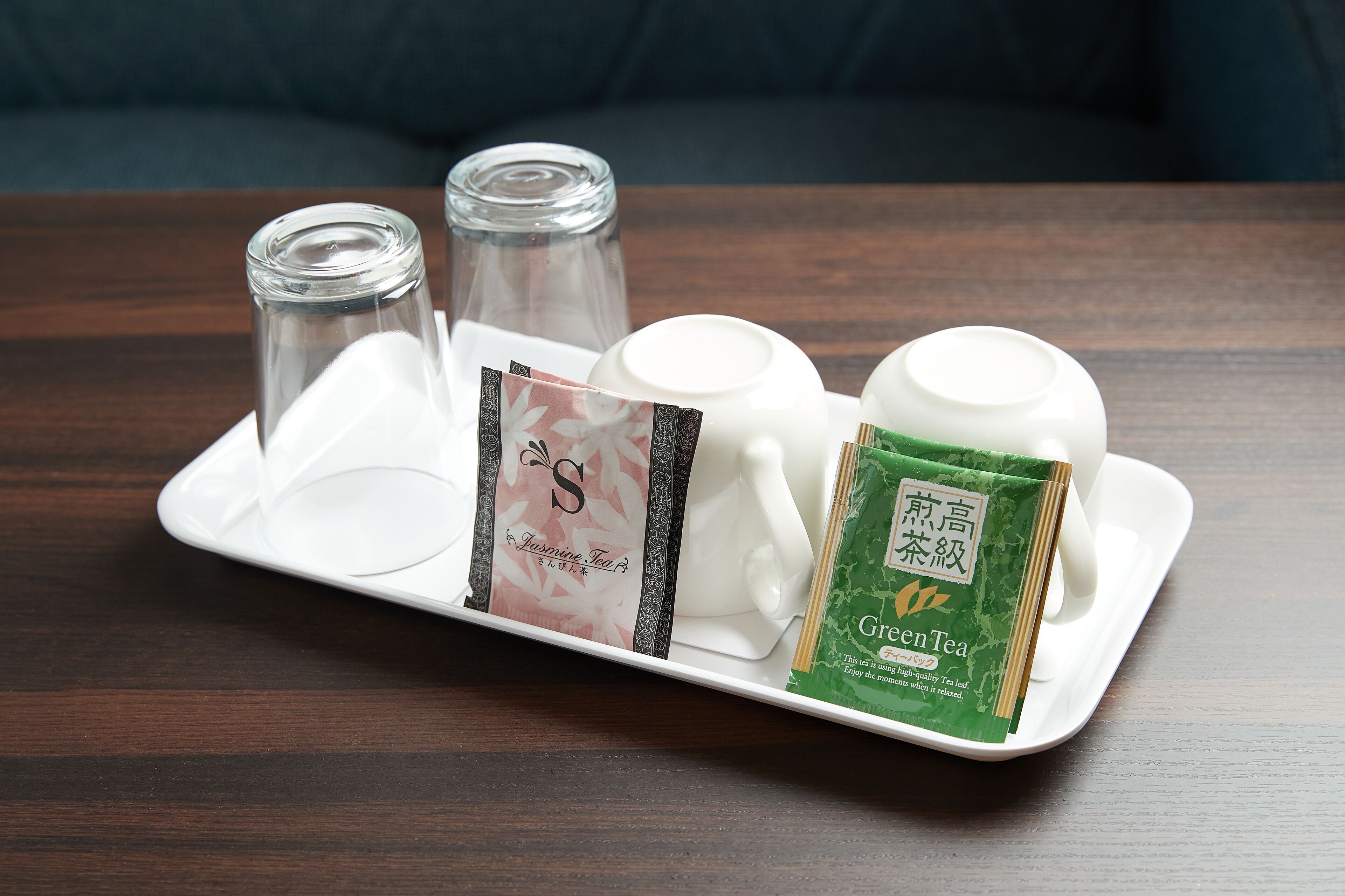 A tray with a white teapot, a glass, two packets of tea, and a small glass jar on a wooden table.