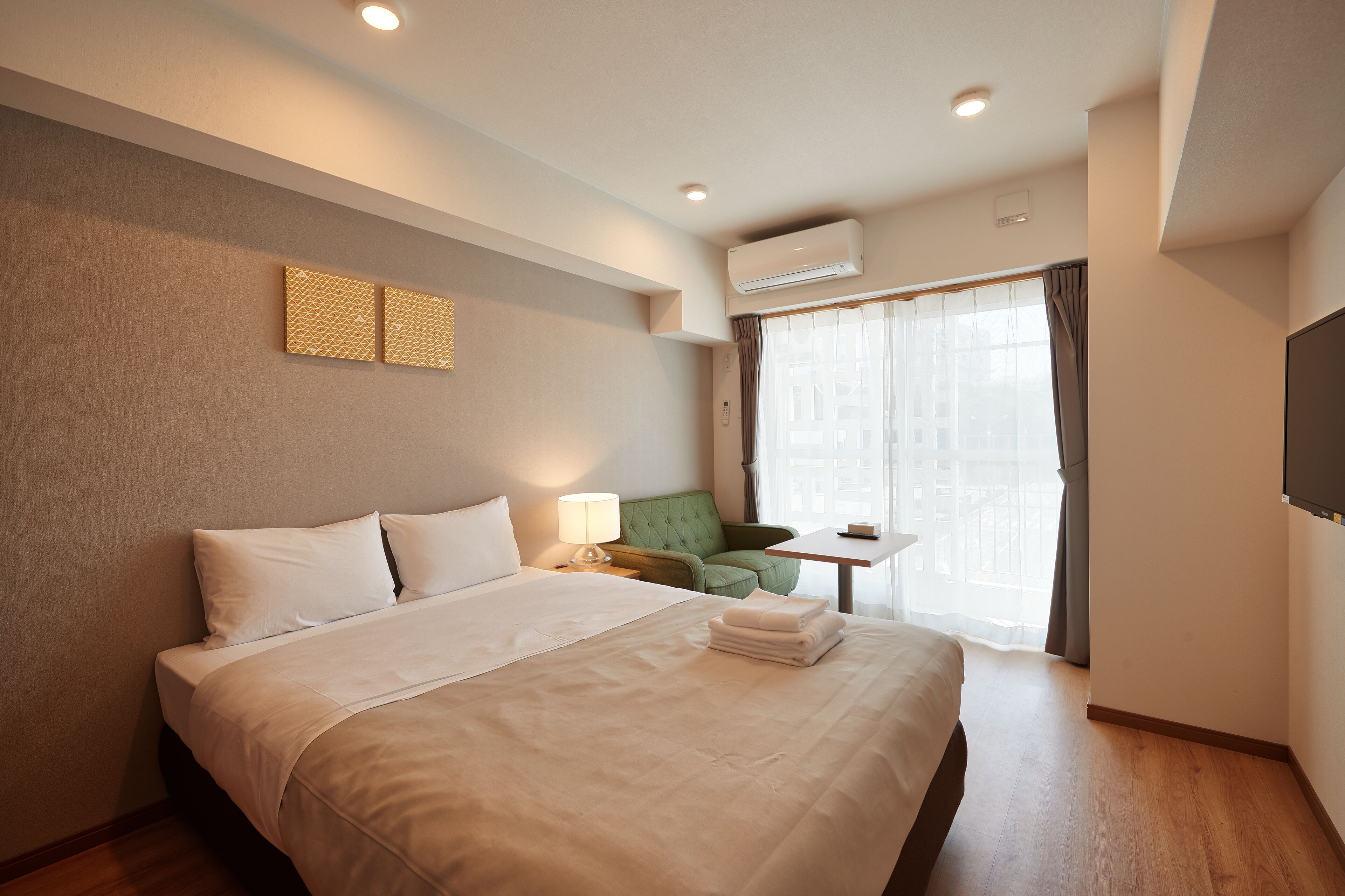 Une chambre d’hôtel avec un lit, un fauteuil vert, une petite table et une fenêtre avec des rideaux.