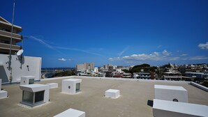 A rooftop with multiple white square structures, a satellite dish, and a view of a cityscape with buildings and a bridge in the distance. - Arce Ryukyu Harbor Stay (Urasoe)