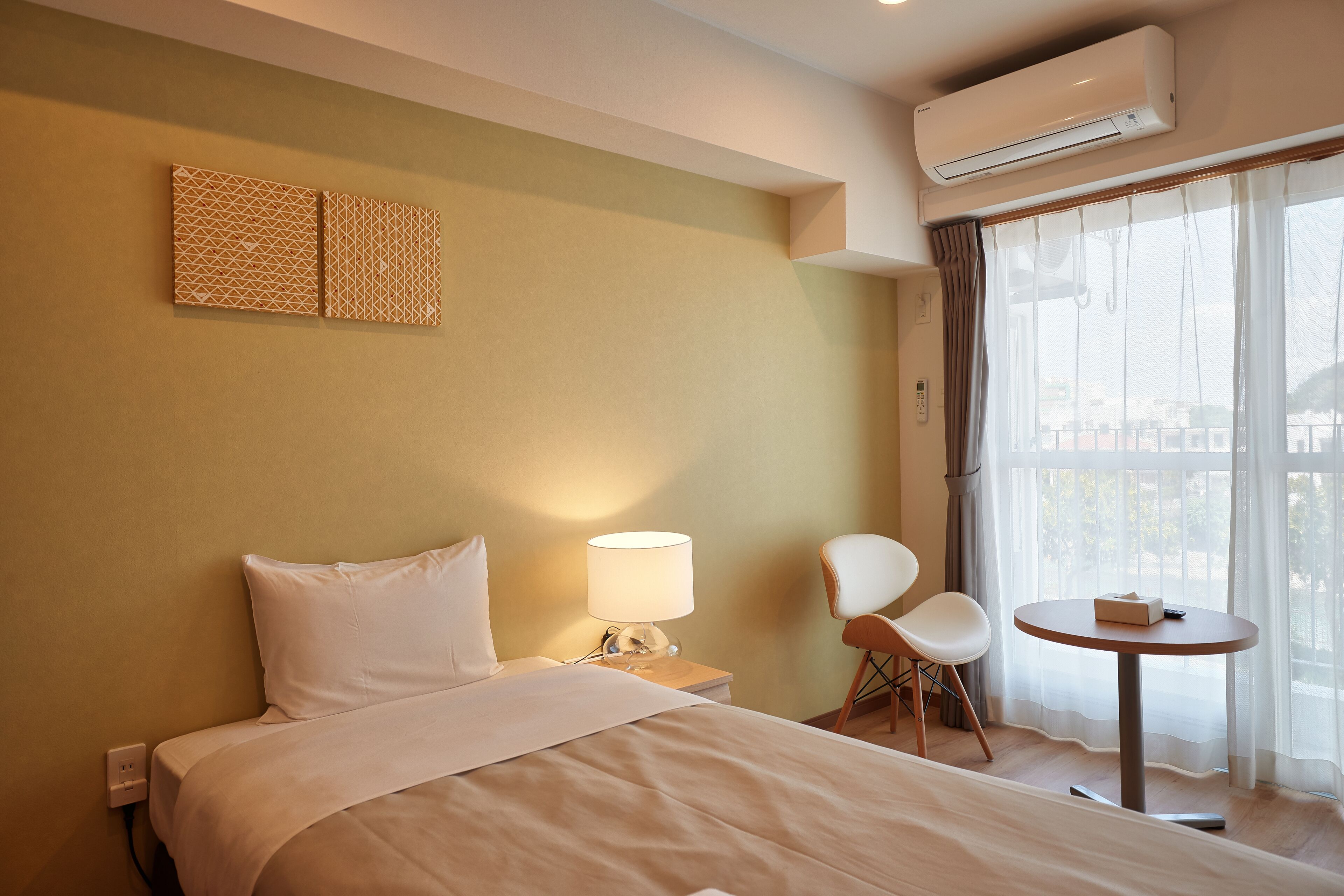 A hotel room with a bed, a bedside table with a lamp, a chair, a small table, and a window with curtains.