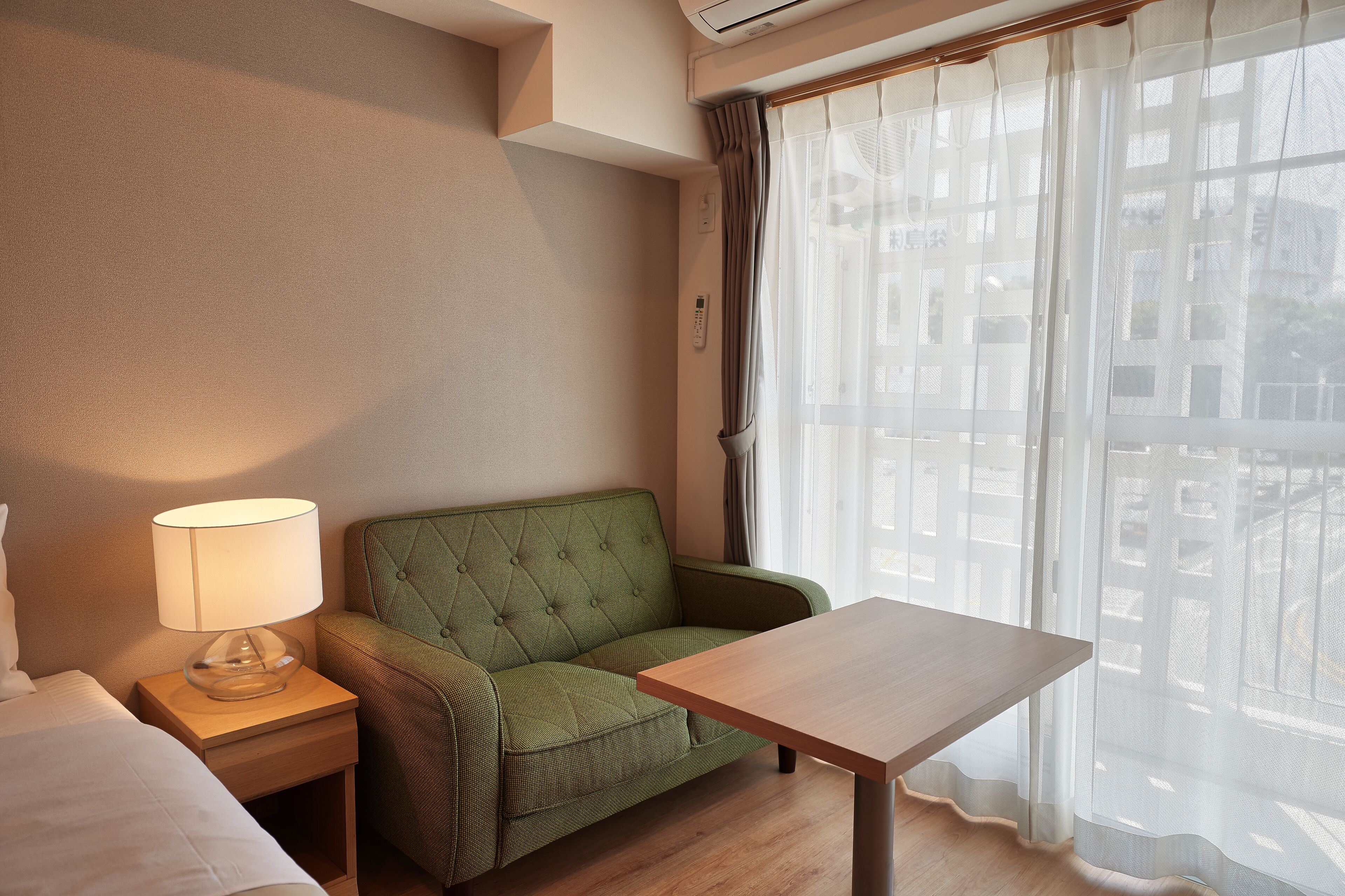 A hotel room with a green sofa, a wooden table, a lamp, and a window with sheer curtains.