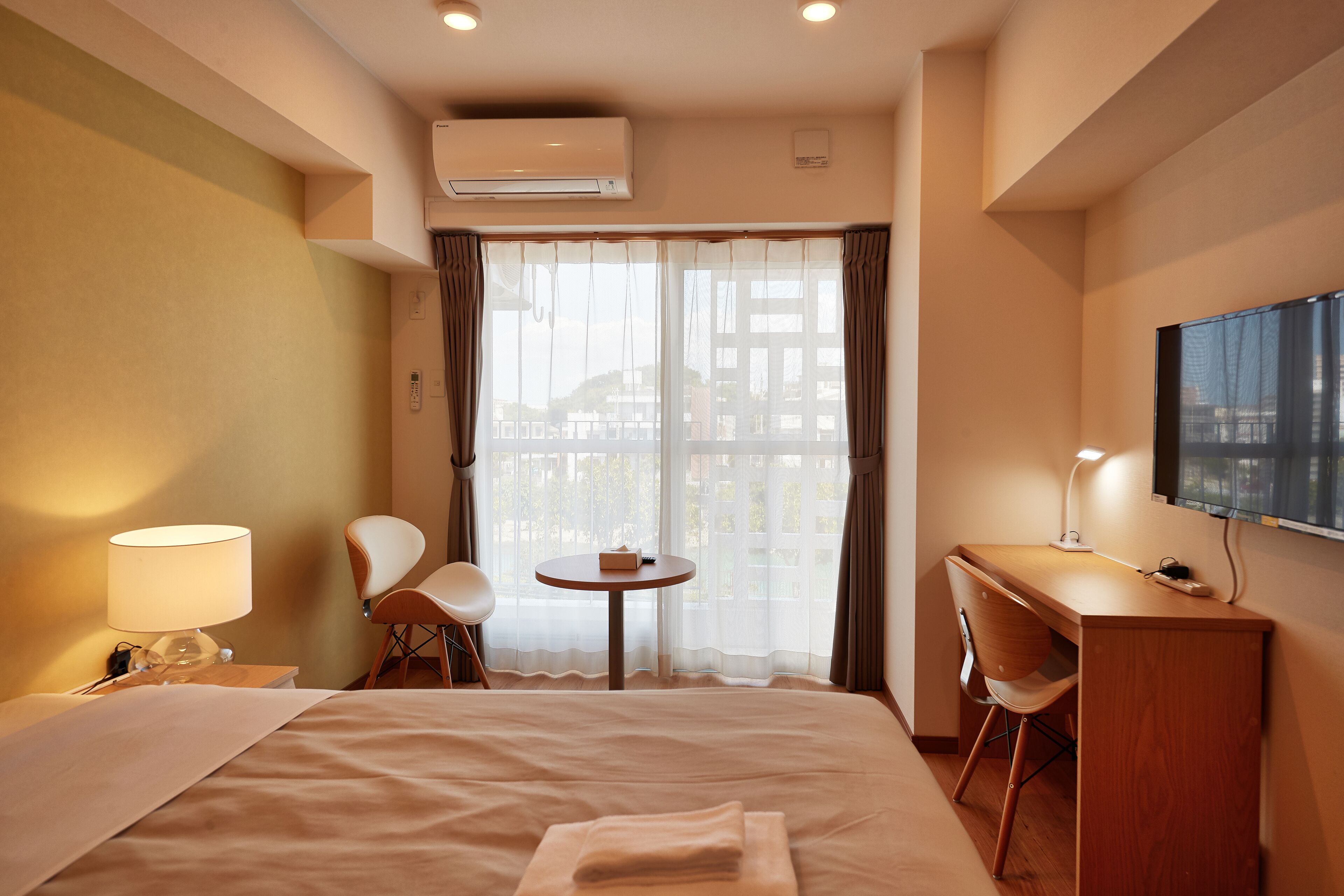 A hotel room with a bed, a desk, a chair, a TV, and an air conditioning unit.