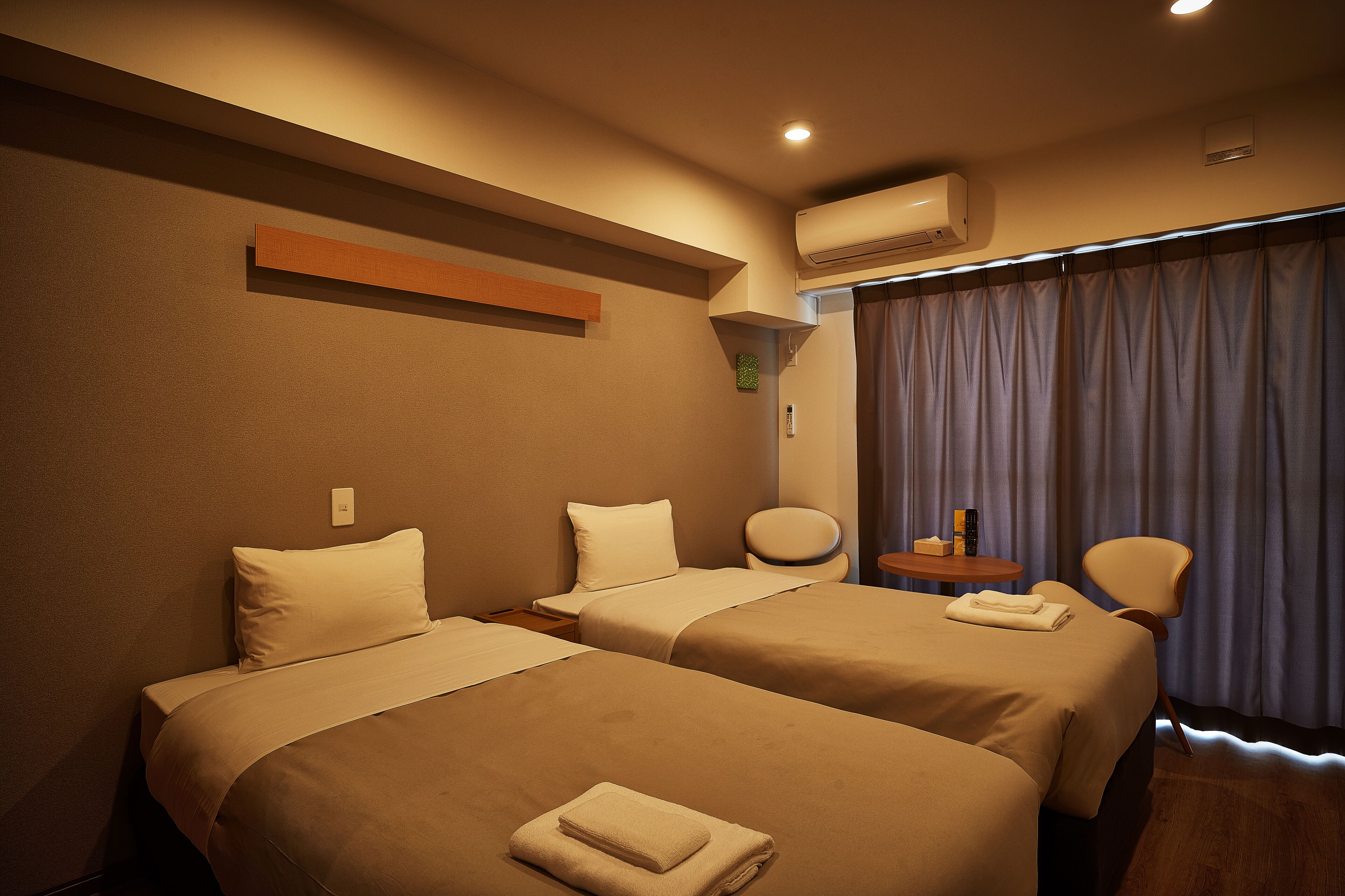 A hotel room with two beds, a round table with a chair, an air conditioning unit, and a window with curtains.