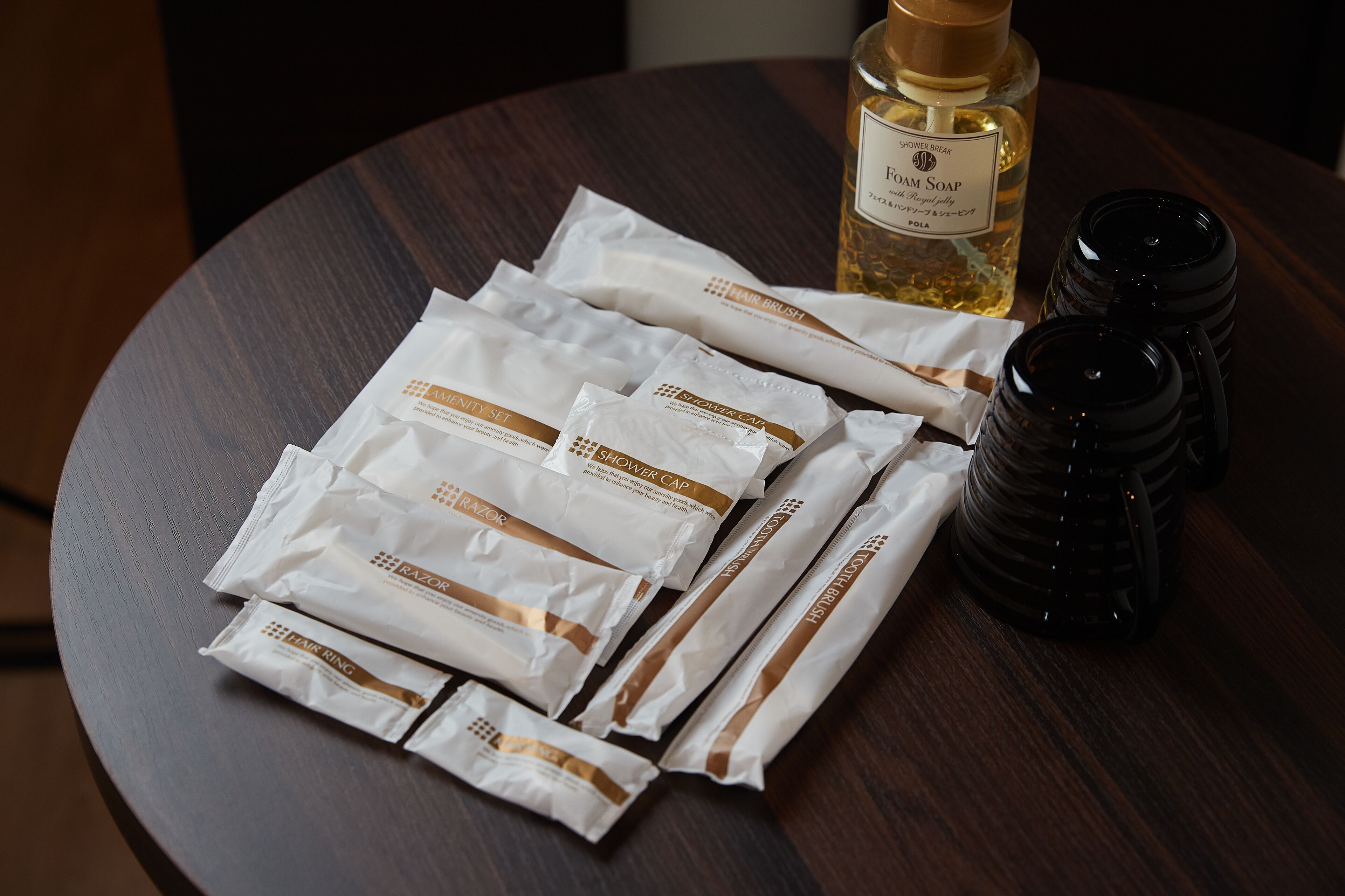 A wooden table with several white and brown packages labeled "Shower Cap" and "Shower Brush," a bottle of "Shower Soap," and a black container.