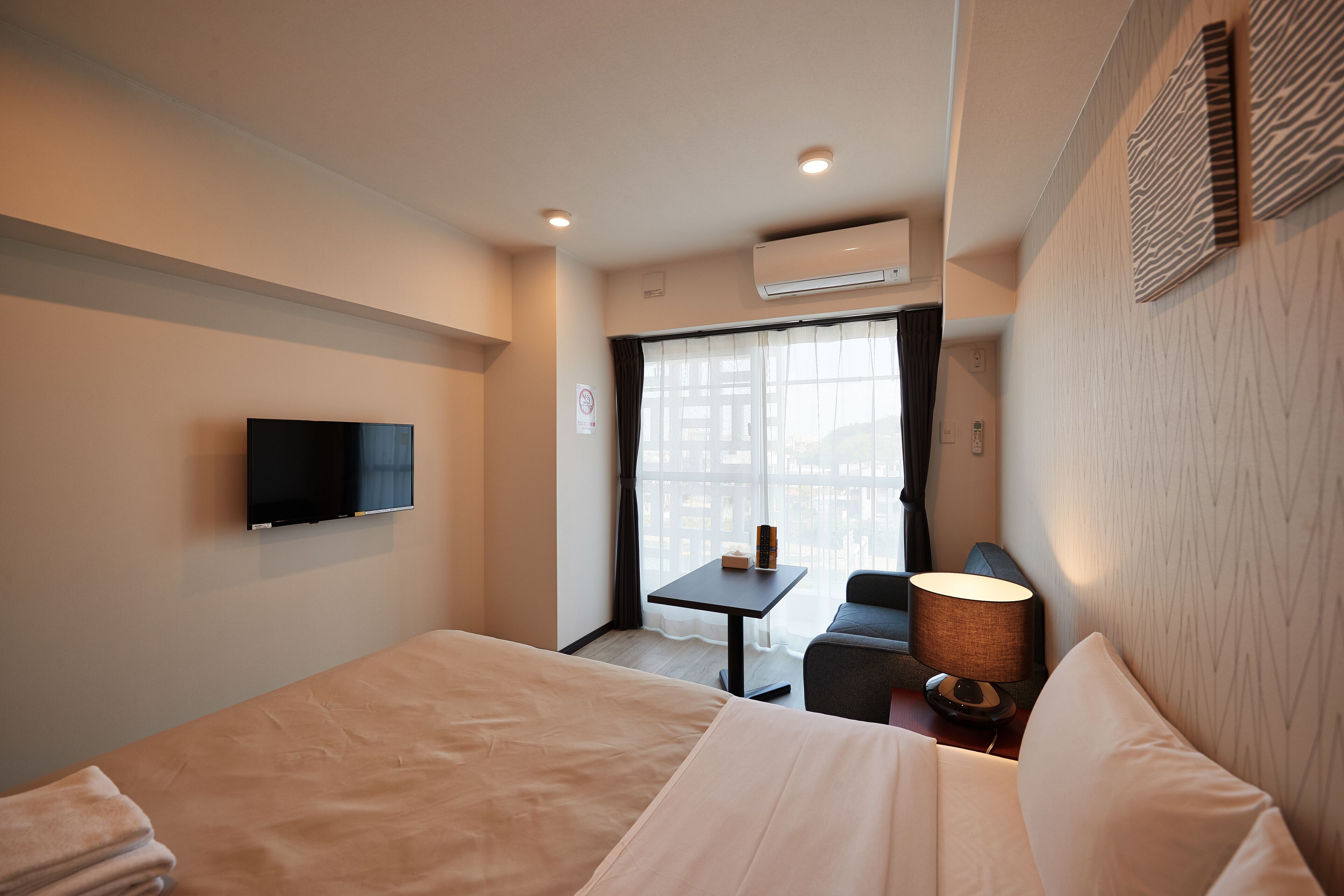 A hotel room with a bed, a TV mounted on the wall, a small table with a lamp, and a view of the city through a window.