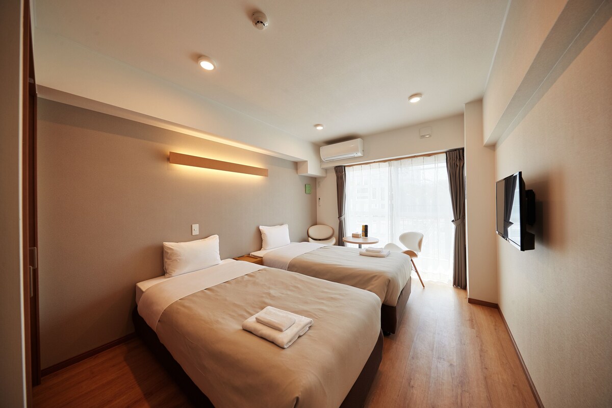 A hotel room with two beds, a TV, a small table, and a window with curtains.