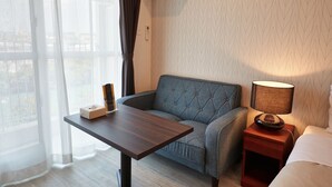 A hotel room with a sofa, a small table, a bedside table with a lamp, and a window with sheer curtains. - Arce Ryukyu Harbor Stay (Urasoe)