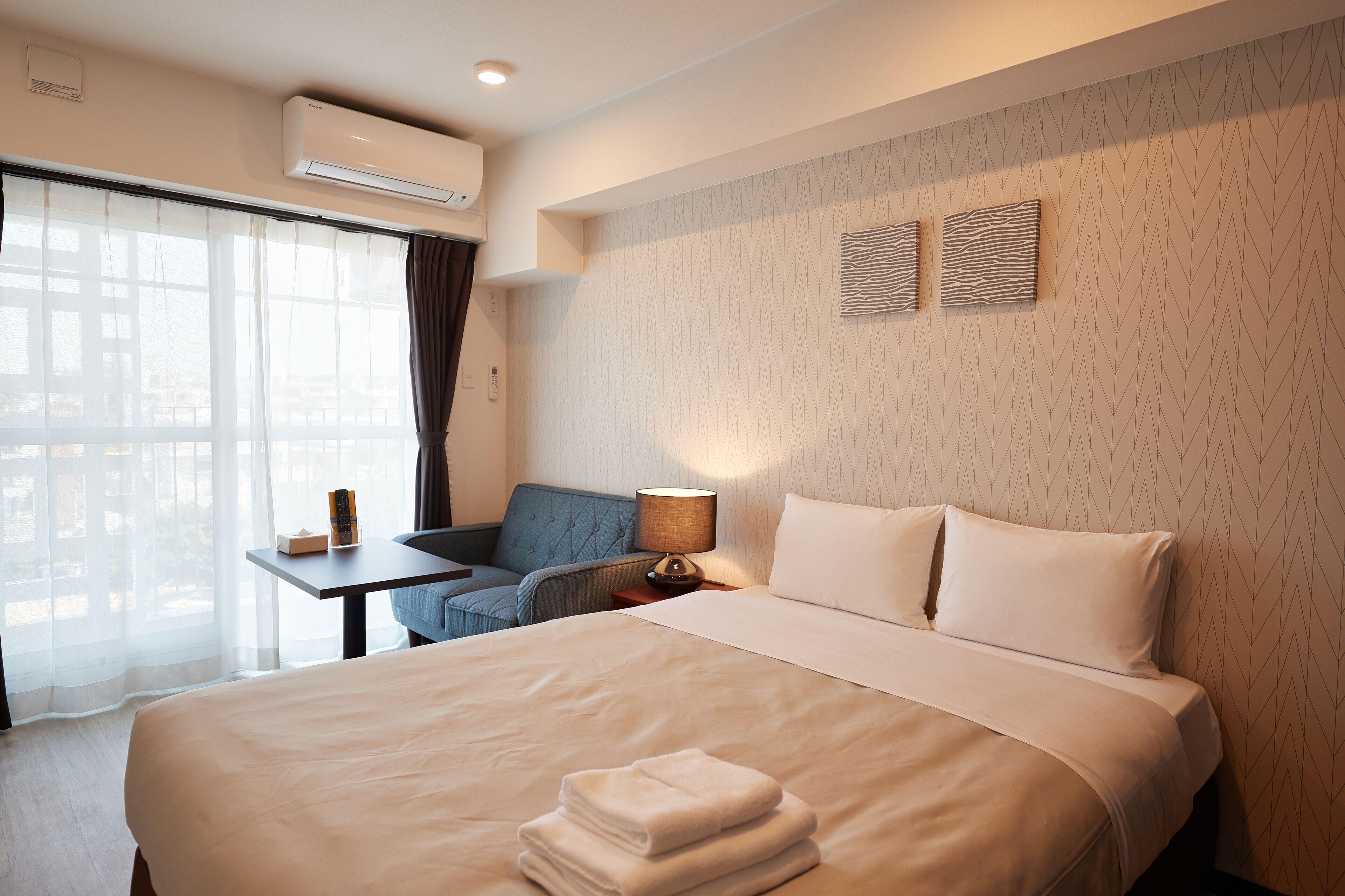 A hotel room with a large bed, a desk with a lamp, a chair, and a view of the city through the window.