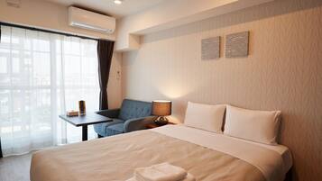 A hotel room with a large bed, a desk with a lamp, a chair, and a view of the city through the window.