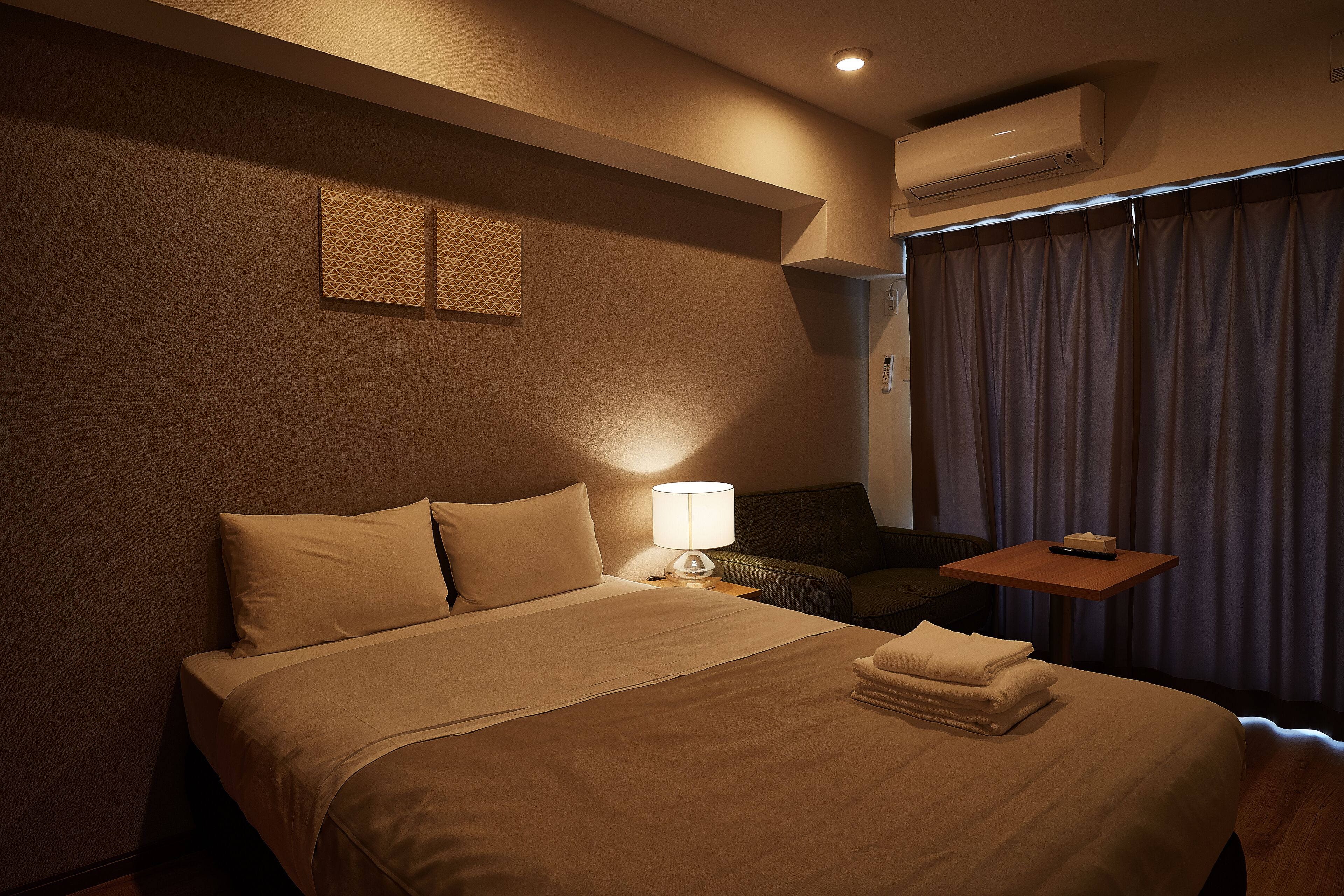 Habitación de hotel con cama, mesita de noche con lámpara, silla y ventana con cortinas.