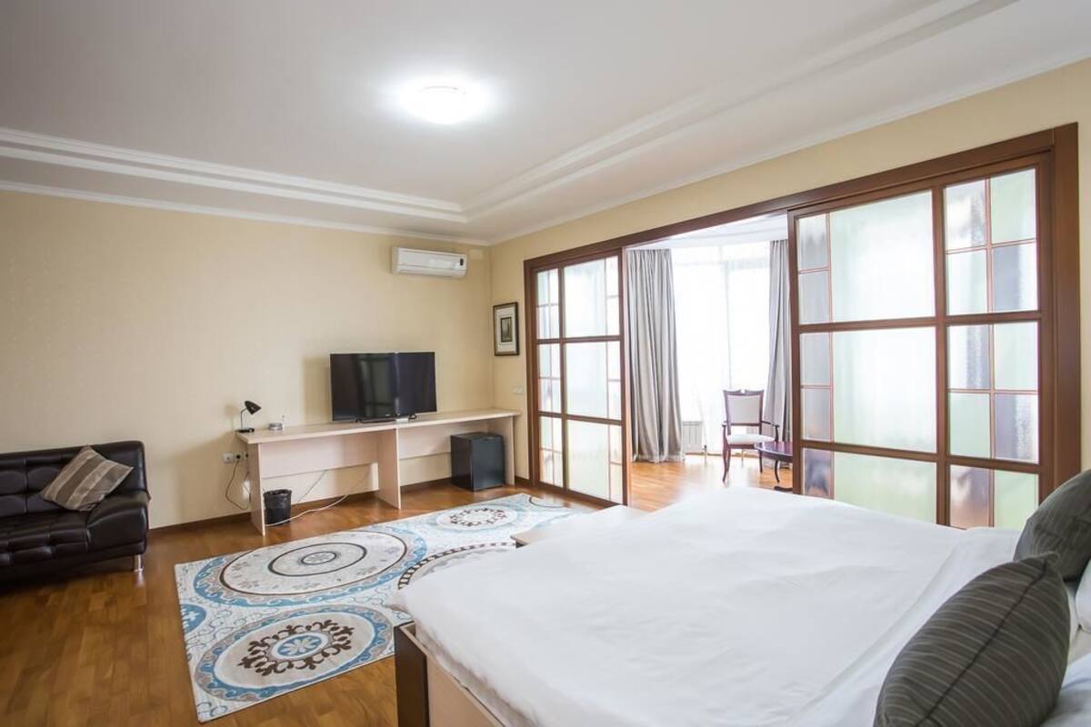 A bedroom with a bed, a desk with a computer, a TV, and a window with curtains.