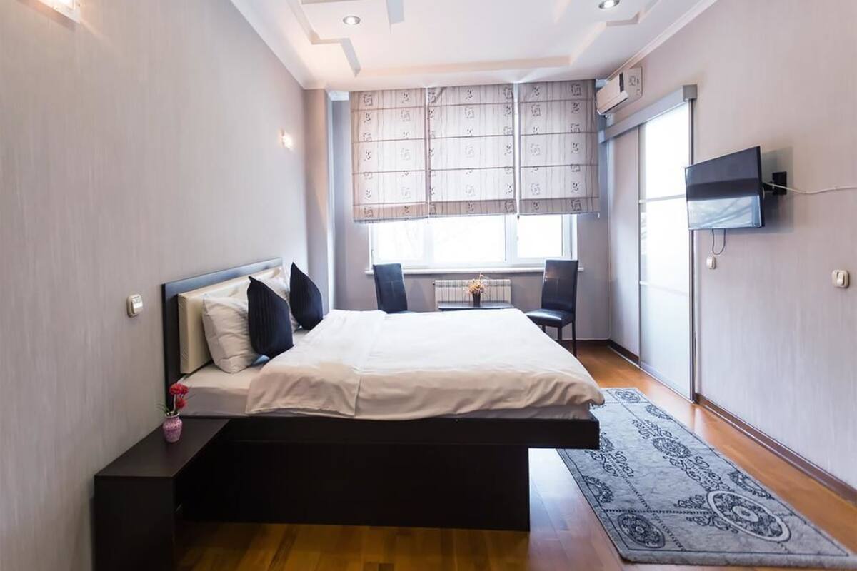A modern bedroom with a bed, a TV, a window with blinds, and a small table with a vase.