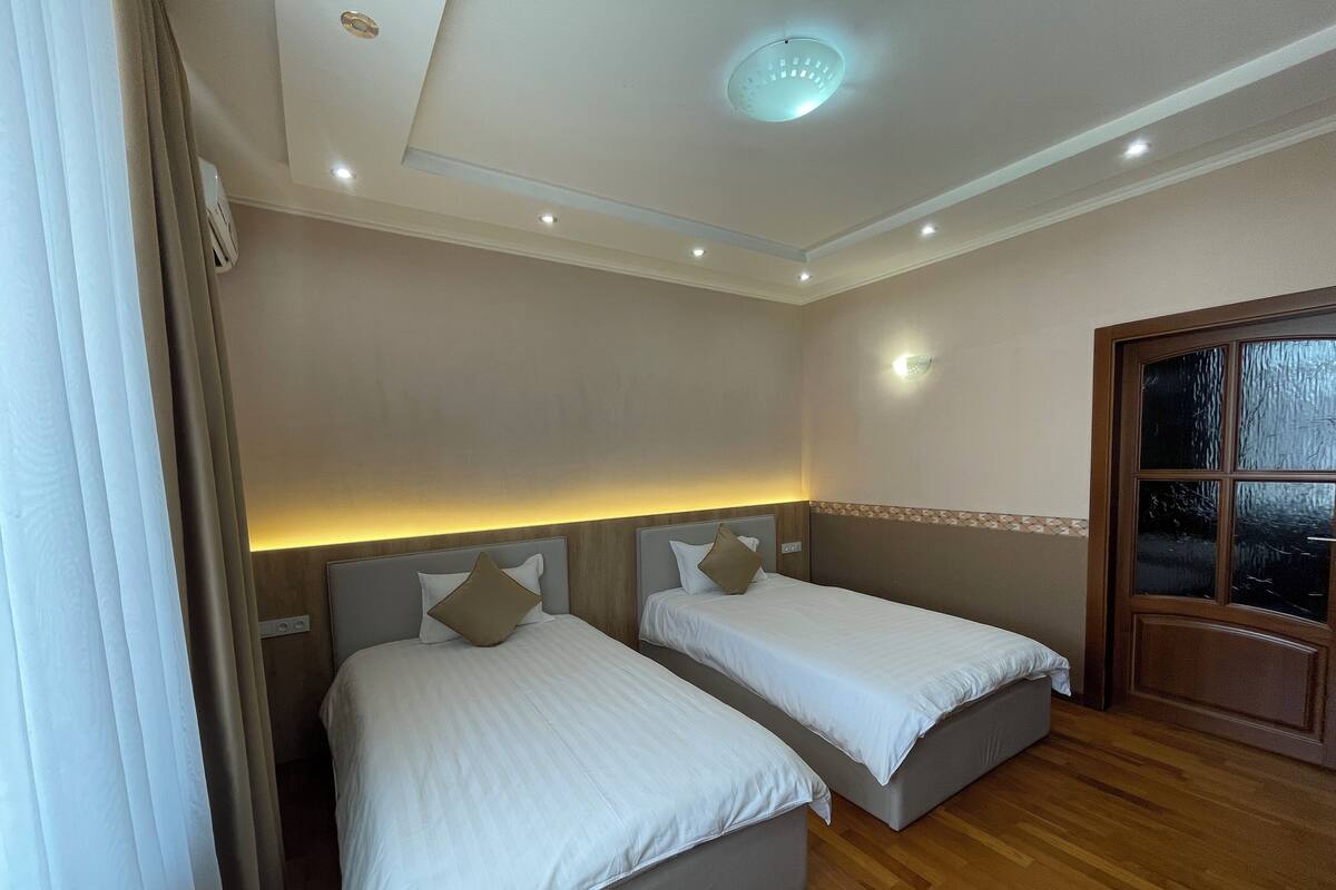 A hotel room with two single beds, wooden flooring, a ceiling light, and a window with a view.