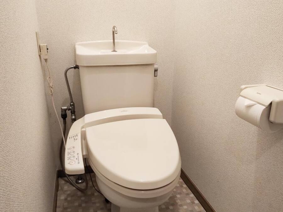 Eine weiße Standardtoilette mit Spülhebel und Bidetanschluss, in einem kleinen Badezimmer mit Fliesenboden und schlichter Wand.
