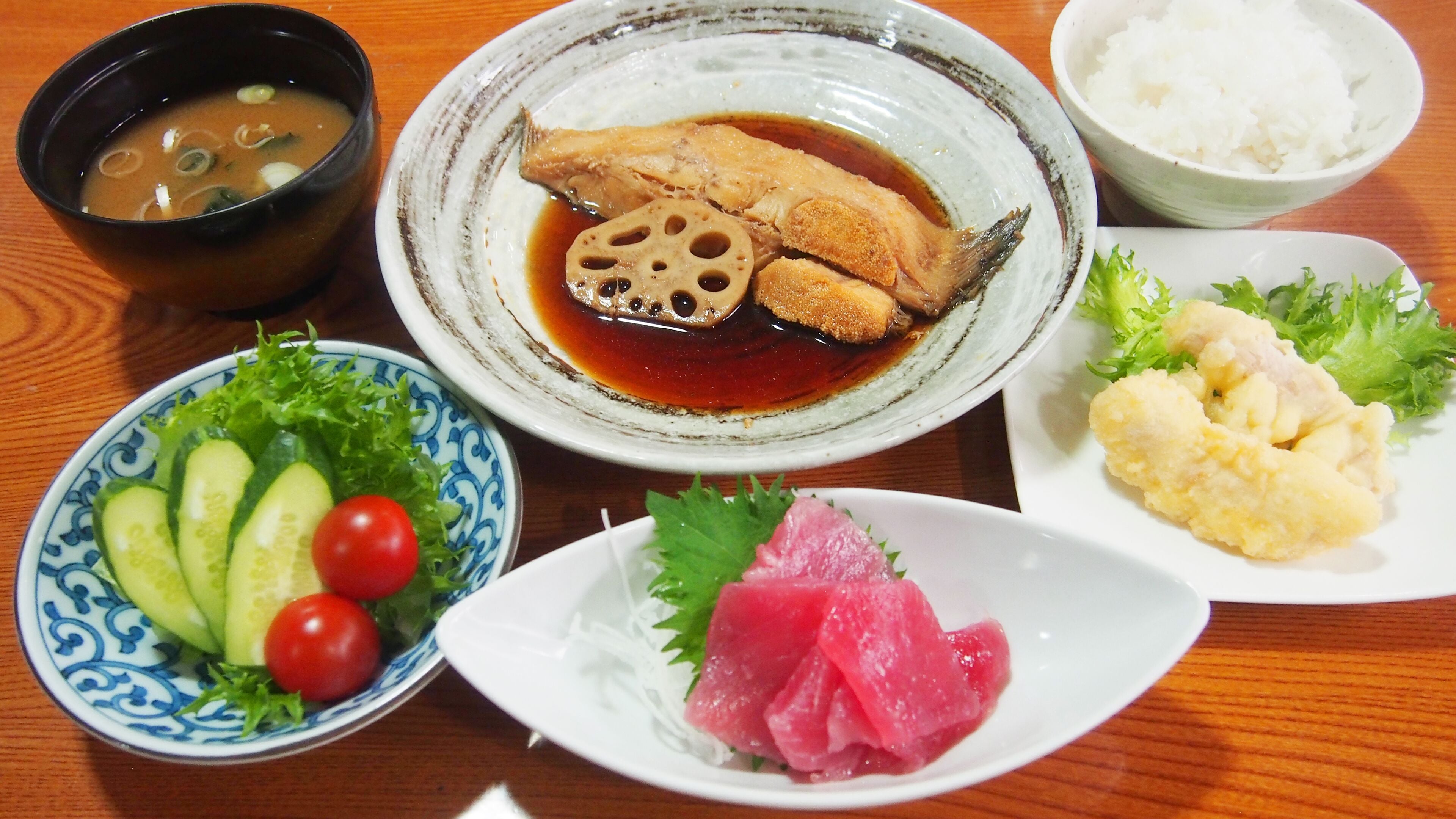 A table with various dishes including a bowl of soup, a plate with fish, a bowl of rice, a plate with cucumber and cherry tomatoes, and a plate with a fish topping.