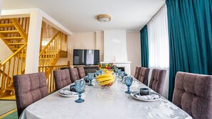 Een eetgedeelte met een lange tafel voor zes personen, omringd door gestoffeerde stoelen. Een televisie is aan de muur bevestigd, en een trap is links te zien.