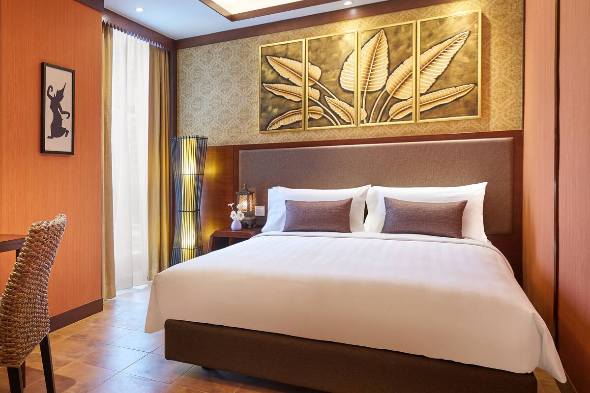 A hotel room with a large bed, a decorative wall panel, a standing lamp, and a wicker chair.