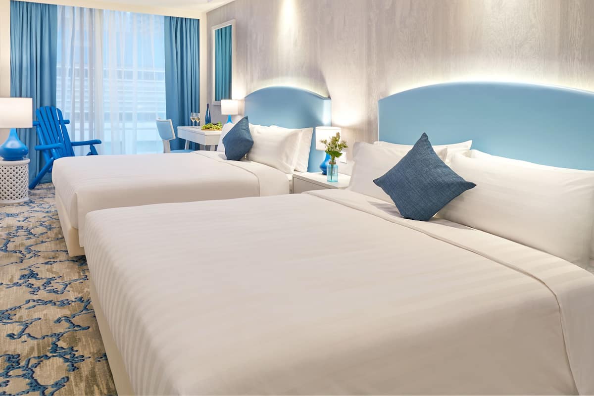 A hotel room with two beds, a desk, a chair, and a view of the sky through the ceiling.
