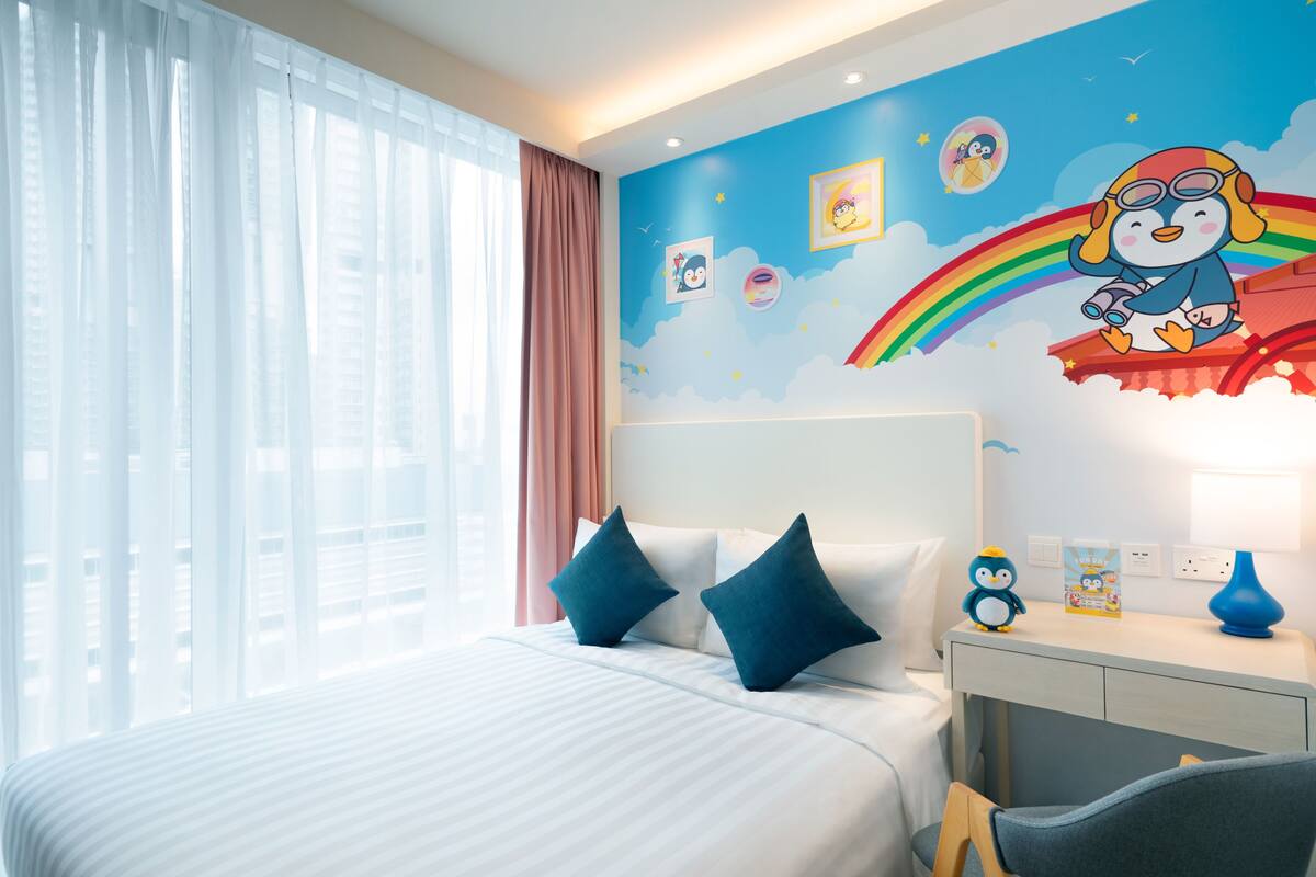 A hotel room with a large bed, two pillows, a desk with a lamp, and a wall with a colorful mural.