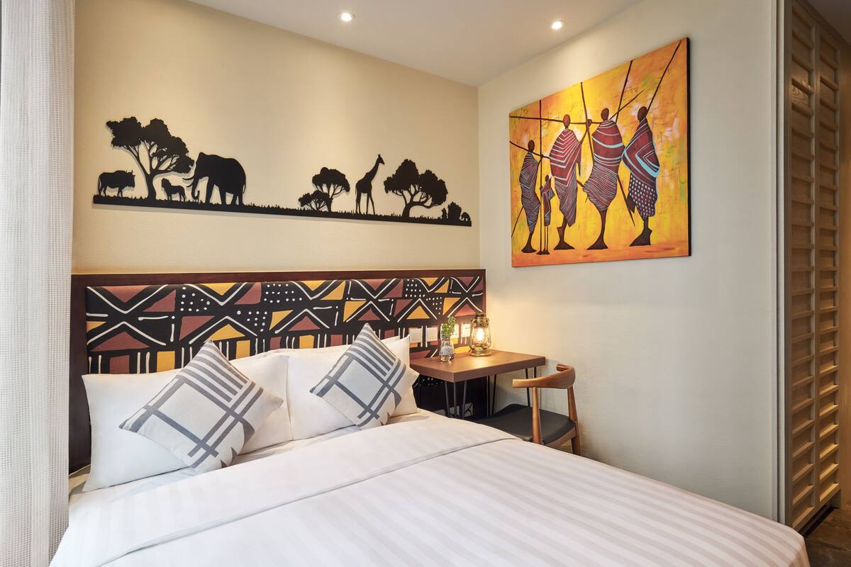 A bedroom with a bed, pillows, a headboard with a geometric pattern, a table with a lamp, and two wall art pieces depicting wildlife and people.