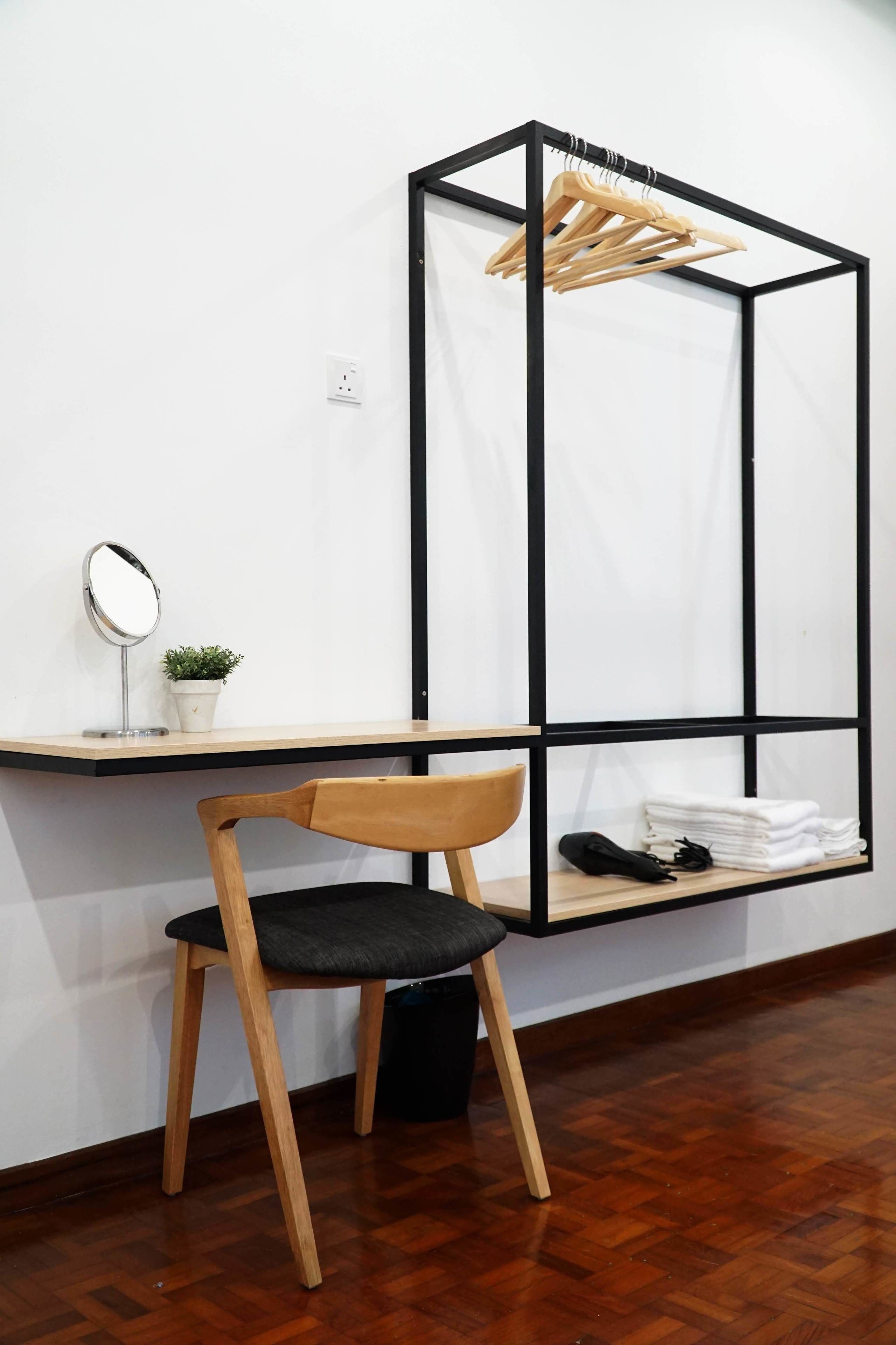 A room with a wooden chair, a small table with a mirror and a potted plant, a black-framed hanging clothes rack, and a shelf with folded clothes and shoes.
