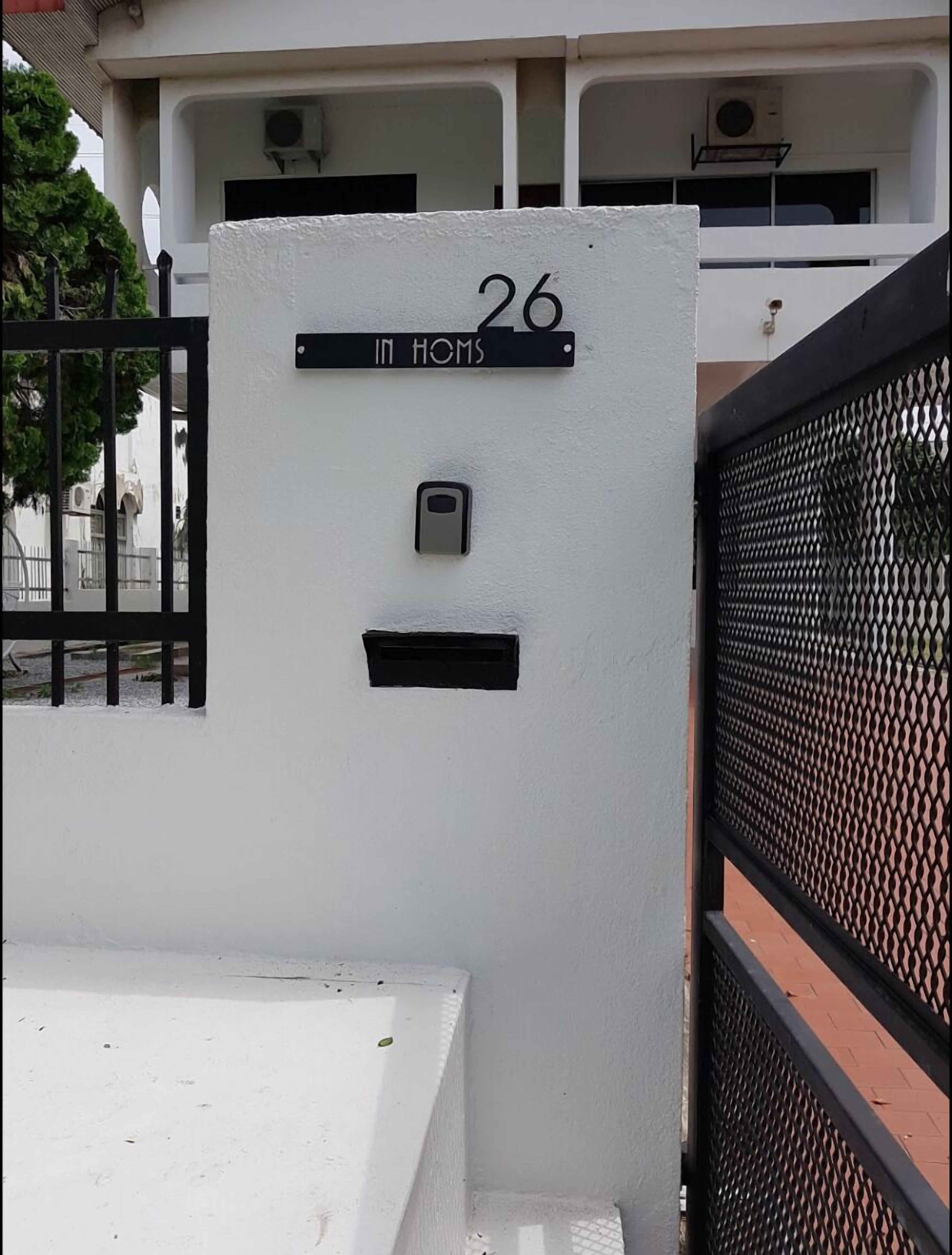 A white building with a black gate, a mailbox, and a sign reading "26 IN HOMS".