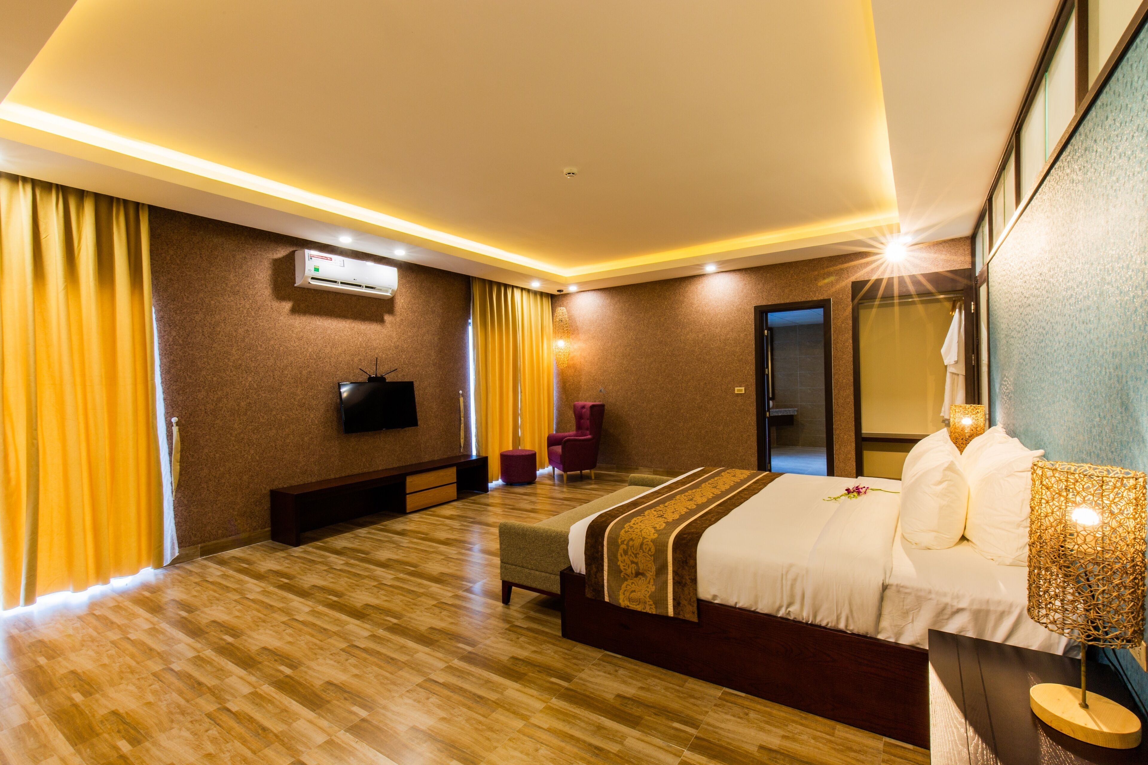 A hotel room with a wooden floor, a bed with a white and brown bedspread, a bench, a TV mounted on the wall, and a window with yellow curtains.