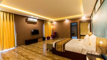 A hotel room with a wooden floor, a bed with a white and brown bedspread, a bench, a TV mounted on the wall, and a window with yellow curtains.