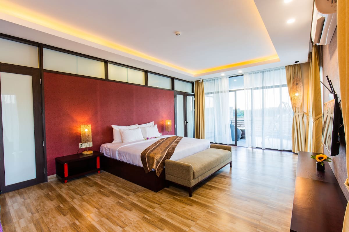 Une chambre d’hôtel moderne dotée d’un grand lit, d’une table de chevet avec une lampe, d’une vue sur l’extérieur grâce à de grandes fenêtres et d’une porte-fenêtre coulissante donnant sur un balcon.