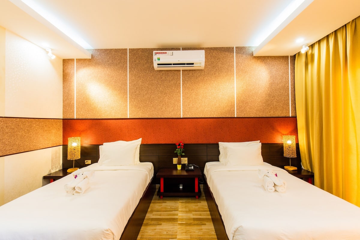 Une chambre d’hôtel avec deux lits, un appareil de climatisation, une table de chevet avec une lampe et une petite table sur laquelle est posé un vase de fleurs.