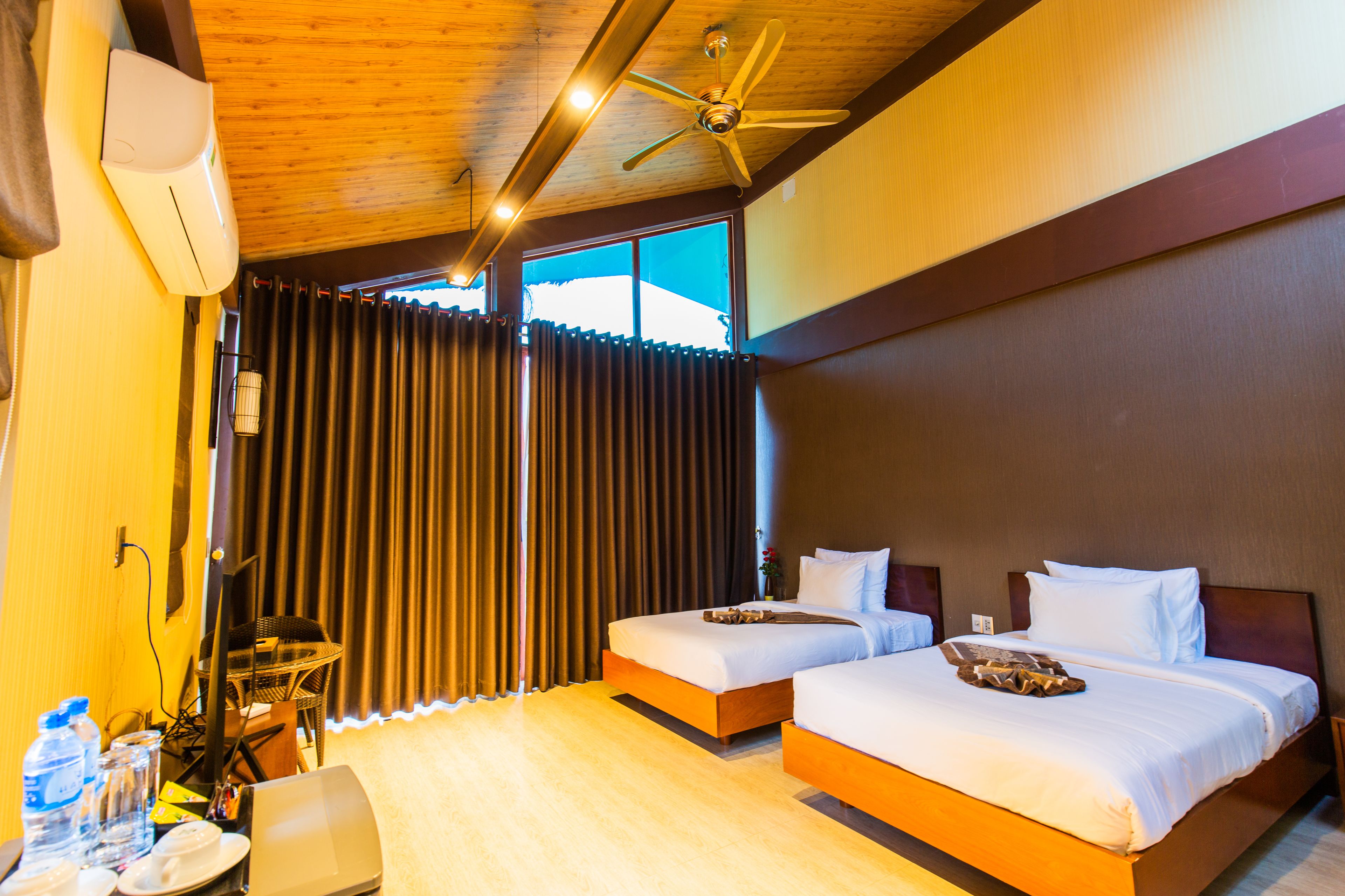 Habitación de hotel con dos camas, ventilador de techo, ventana grande con cortinas y piso de madera.