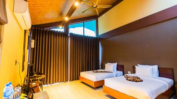 Habitación de hotel con dos camas, ventilador de techo, ventana grande con cortinas y piso de madera.