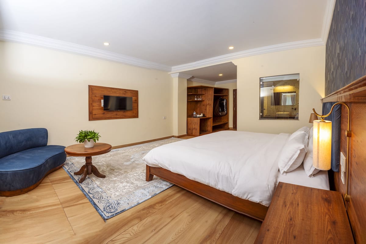 A bedroom with a bed, a TV mounted on the wall, a blue sofa, a small table with a plant, and a wooden nightstand.