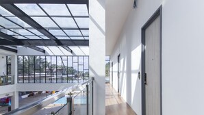 Een moderne hotelgang met een glazen balustrade, een zwembadgedeelte met een oldtimer en een fiets, en een uitzicht op de zee en palmbomen.