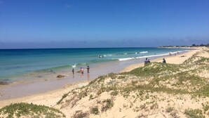 Ein Strand mit klarem, blauem Wasser, Sanddünen und einigen Personen, die spazieren gehen oder stehen.