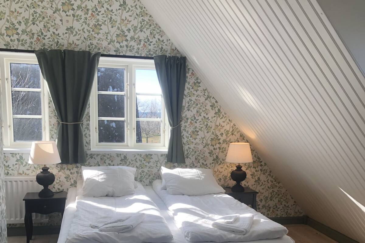 Quarto com uma cama grande, duas mesas de cabeceira com luminárias, uma janela com cortinas e vista para o exterior.