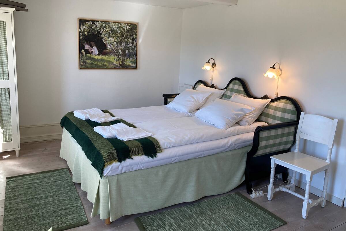 Uma cama bem arrumada com lençóis brancos e cabeceira verde xadrez, uma mesinha de cabeceira, uma cadeira e um quadro na parede.