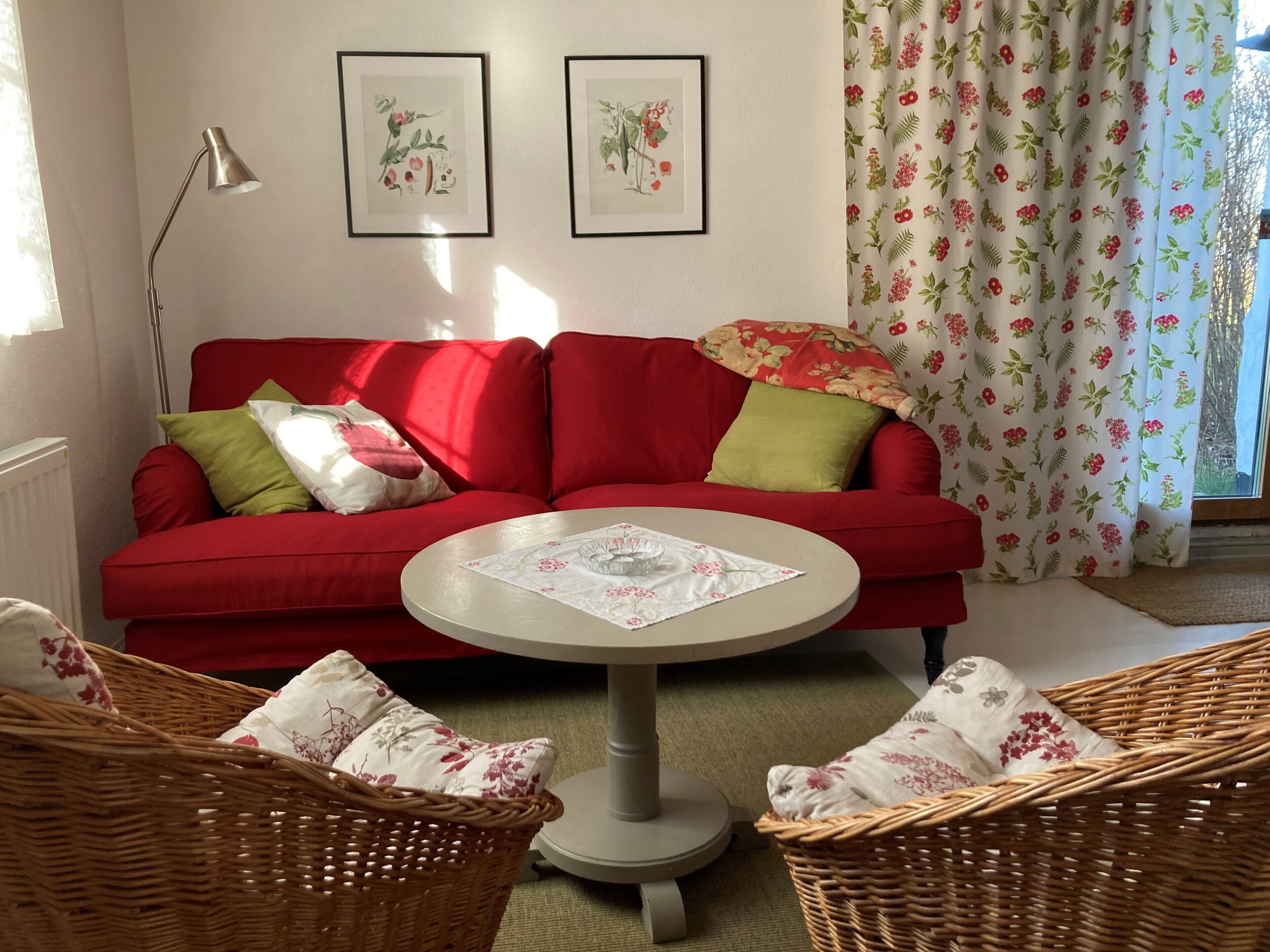 A red sofa with green and white cushions, a round table with a floral tablecloth, wicker chairs with floral cushions, and two framed botanical prints on the wall.