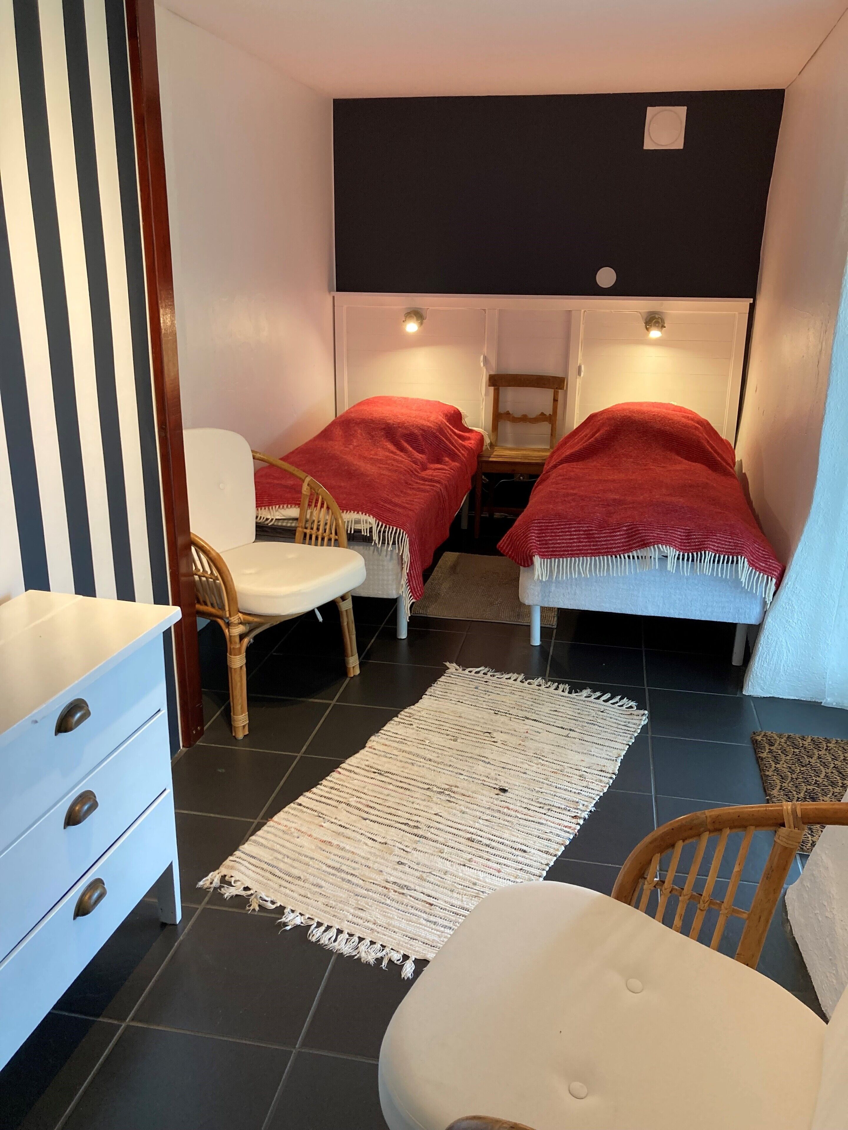Una habitación pequeña y funcional con dos camas individuales, una silla blanca, una silla de madera, un aparador blanco y una alfombra estampada.