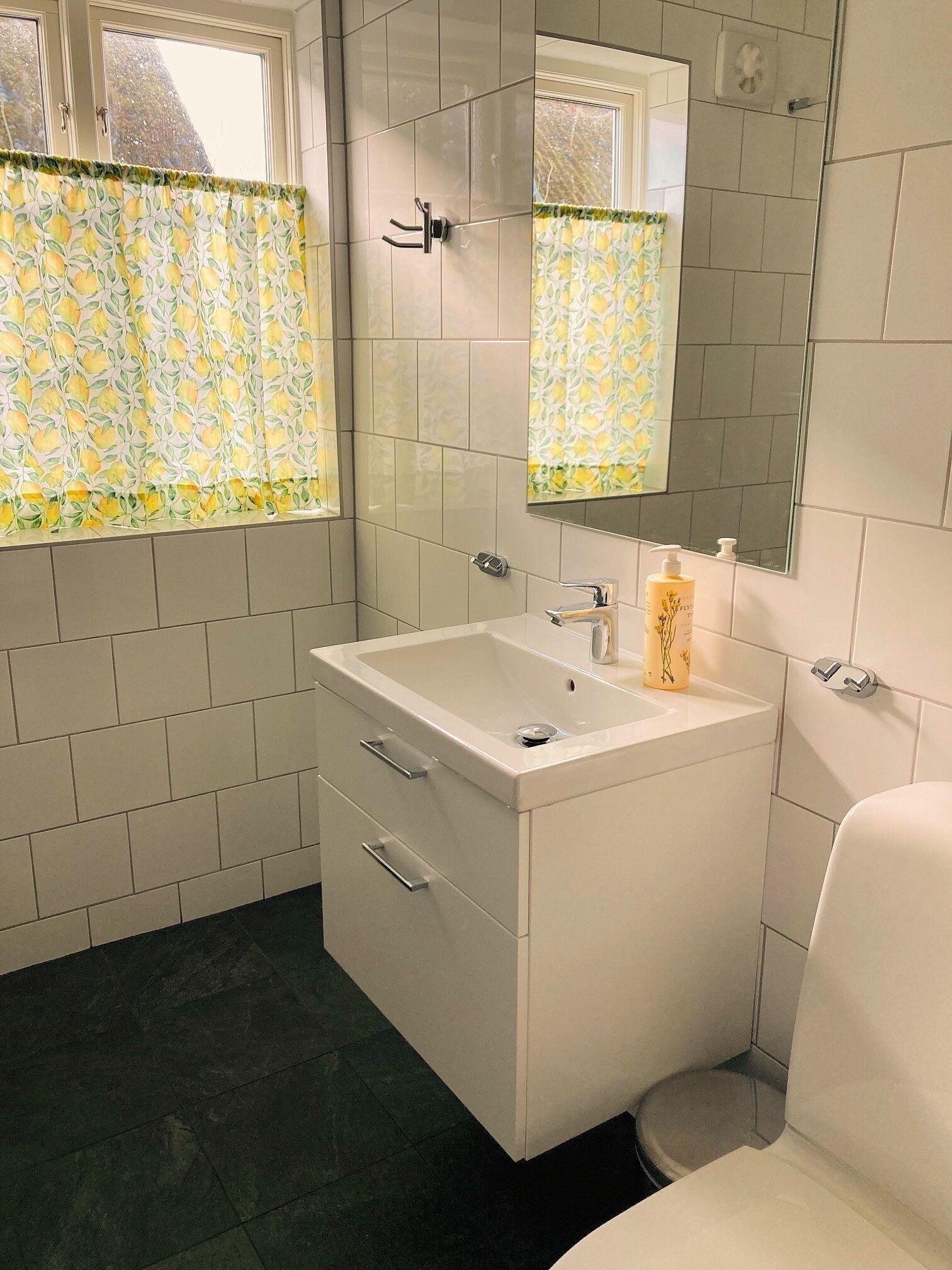 A bathroom with a white sink, mirror, and toilet. There are two windows with lemon-patterned curtains.