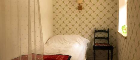 A narrow room with a bed, a chair, a wall-mounted lamp, and a window with a view.