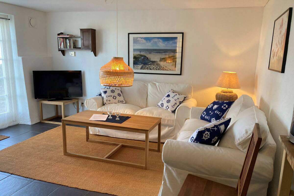 Uma sala de estar com um sofá branco, uma mesa de centro de madeira, uma televisão e um quadro na parede.