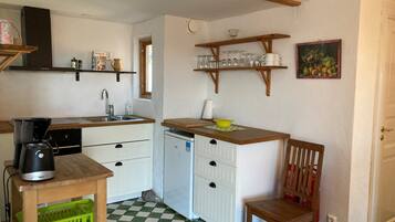 A compact kitchen with a coffee maker, a sink, and a dishwasher. There are wooden shelves above the counter holding dishes and glasses. A chair is placed near the counter, and a painting hangs on the wall.