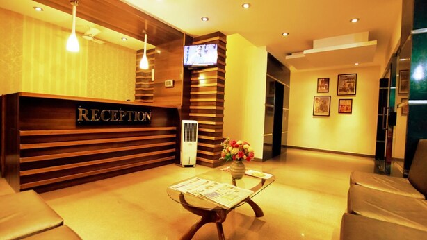 Una zona de recepción con un mostrador de madera, un televisor montado en la pared, un ventilador de pie y un jarrón con flores sobre una mesa.